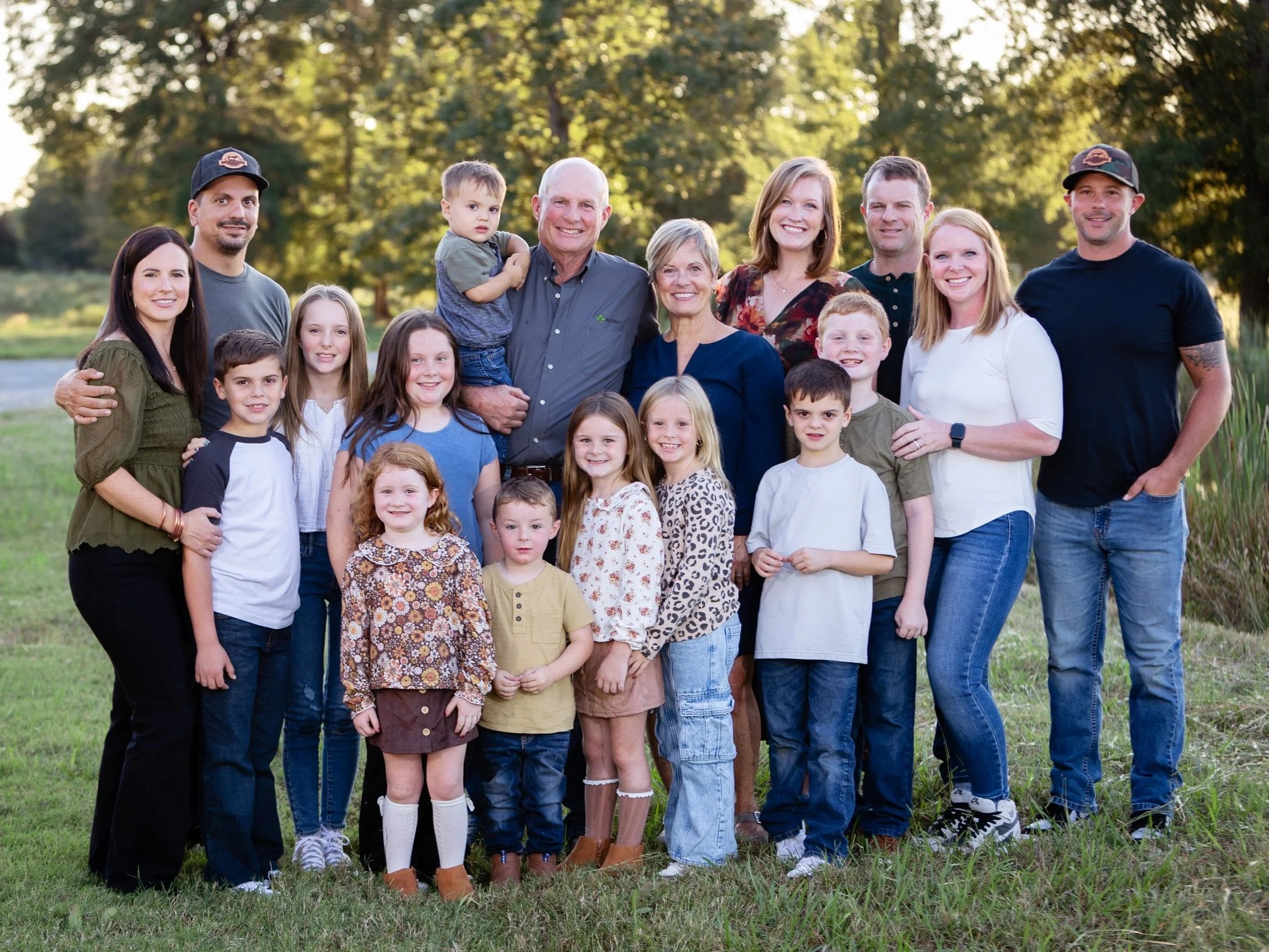 A large family gathering in an outdoor setting, featuring 15 members including adults and children, smiling together in a lush green field with trees in the background. The group showcases a variety of casual clothing styles, reflecting a warm, joyful atmosphere.