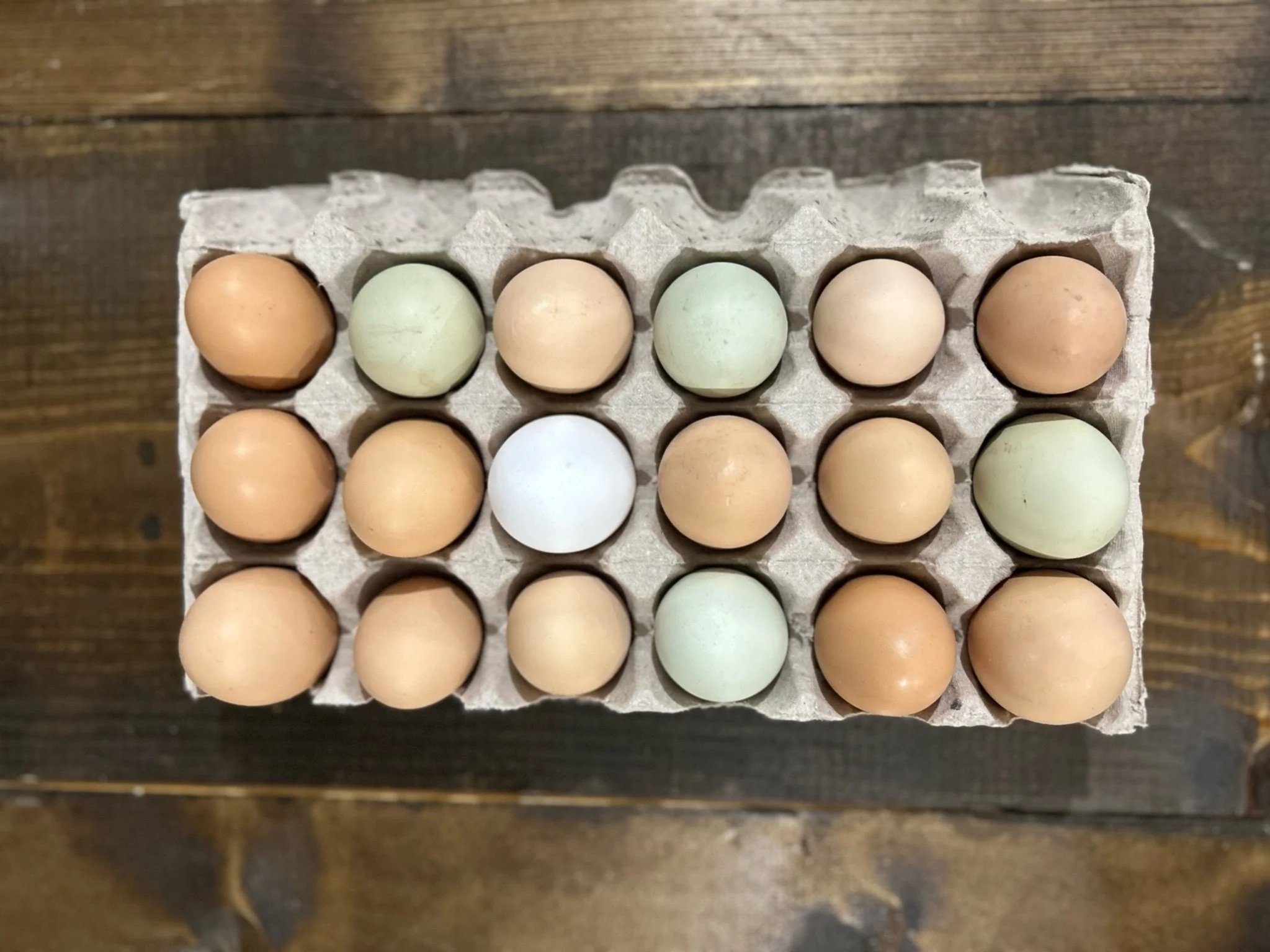 A carton of 18 farm-fresh eggs in various colors, including light brown, green, and white, arranged neatly on a wooden surface.