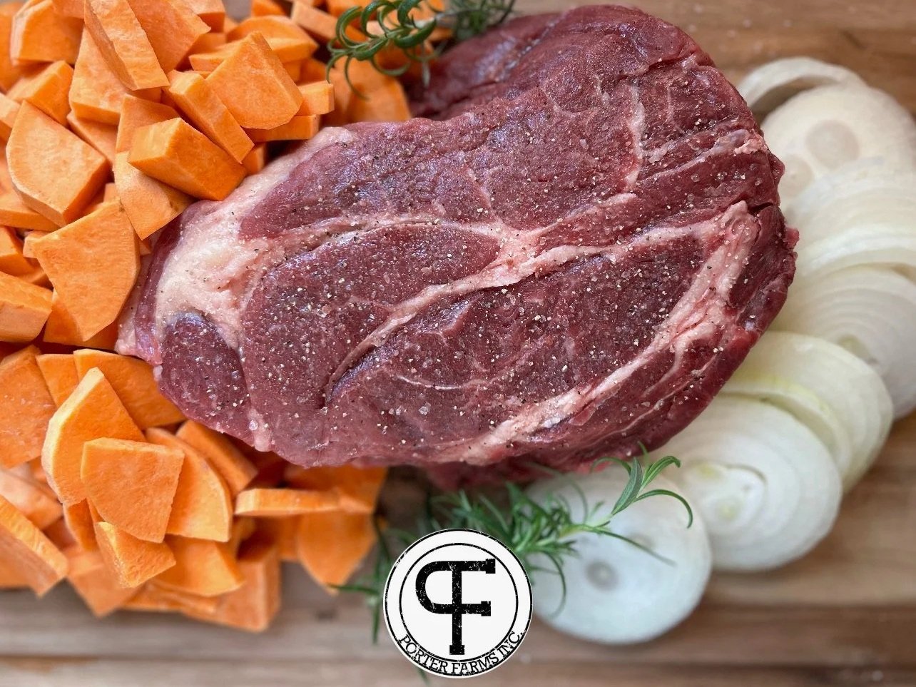 Fresh cut beef roast surrounded by diced sweet potatoes and sliced onions on a wooden cutting board, garnished with rosemary. Ideal for hearty meal preparations and slow cooking recipes.