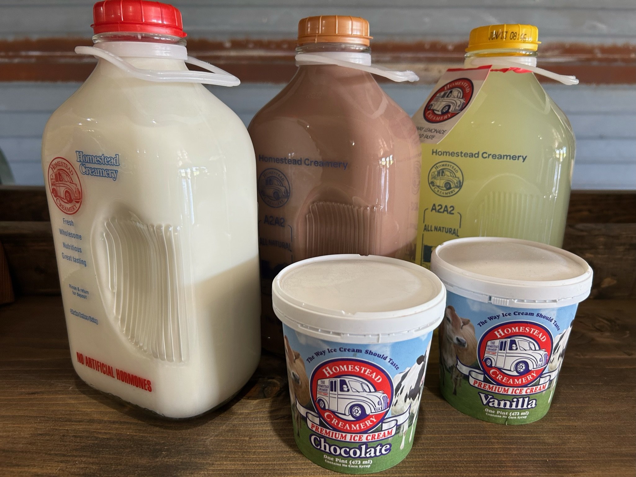 Bottles of Homestead Creamery milk in various flavors, including whole milk and A2A2 milk, alongside containers of chocolate and vanilla ice cream. All products are labeled as all-natural and free from artificial hormones, displayed on a wooden surface.