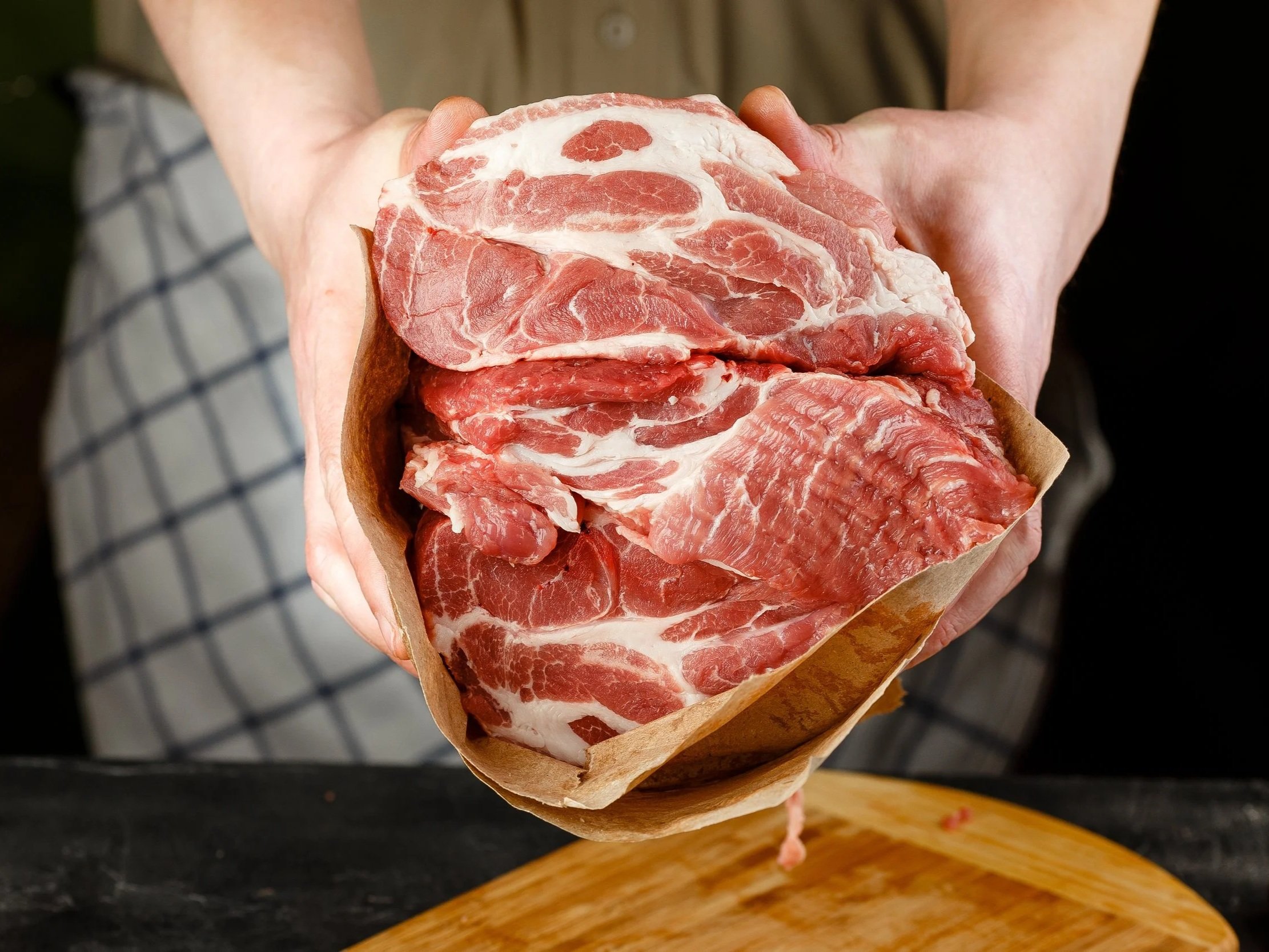 Fresh cuts of pork shoulder wrapped in butcher paper, held by a person in a kitchen setting. The image showcases the marbled texture of the meat, highlighting its quality for cooking and meal preparation.