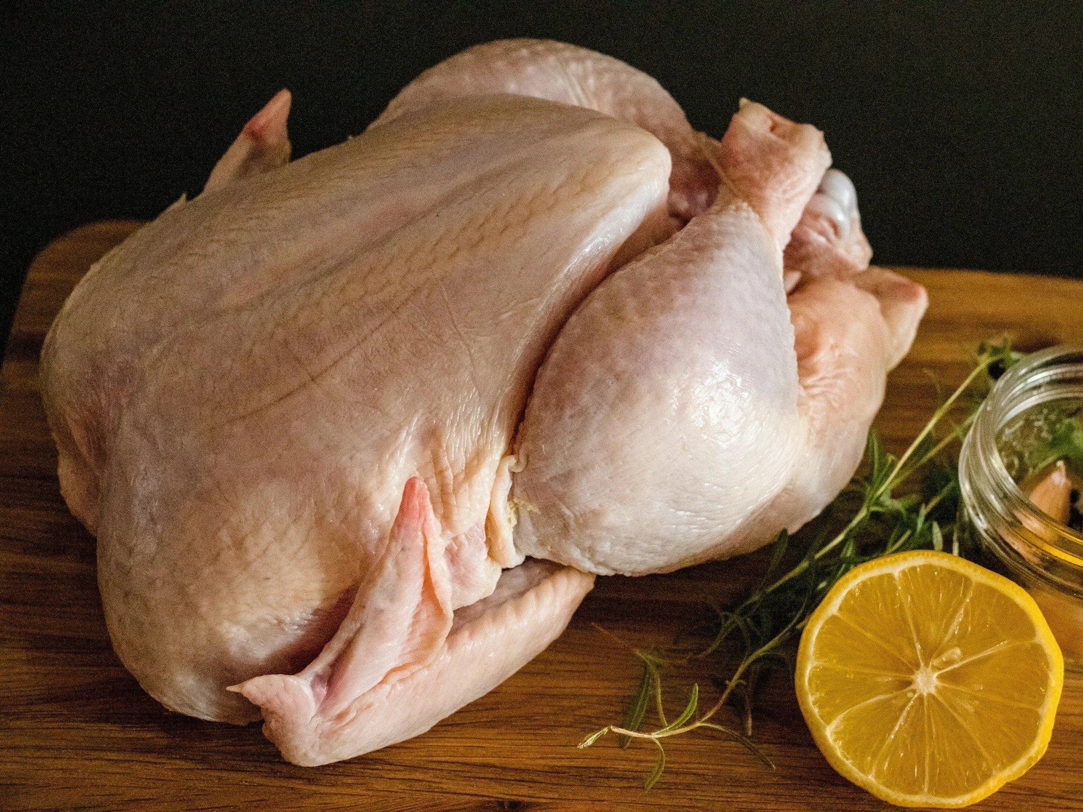 Raw whole chicken on a wooden cutting board, accompanied by fresh herbs and a sliced lemon, ideal for cooking and meal preparation.