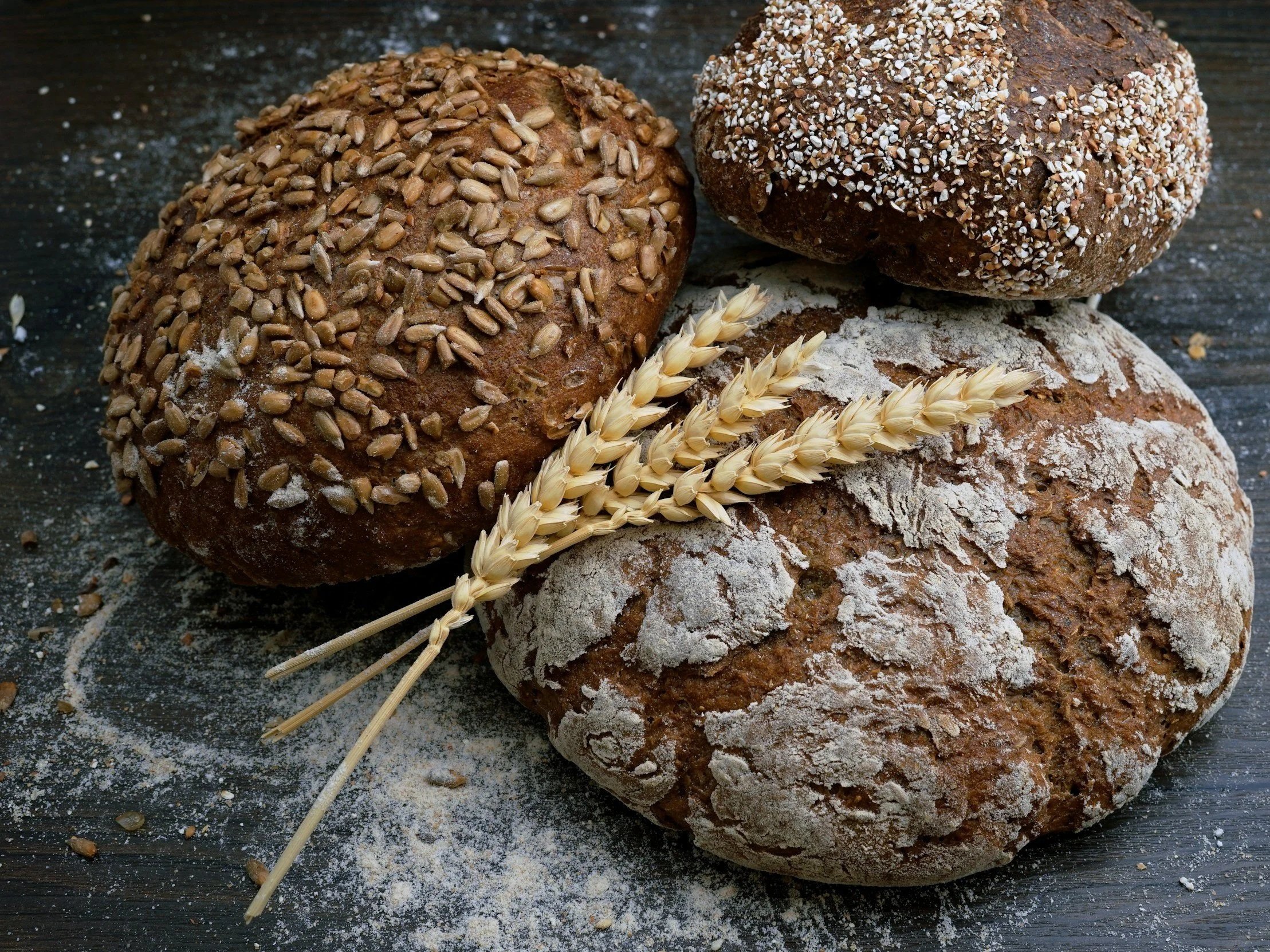 Three types of artisanal bread, including a seeded round loaf, a crusty bread with a floury surface, and a dark bread, arranged with a bundle of wheat stalks on a rustic wooden surface.
